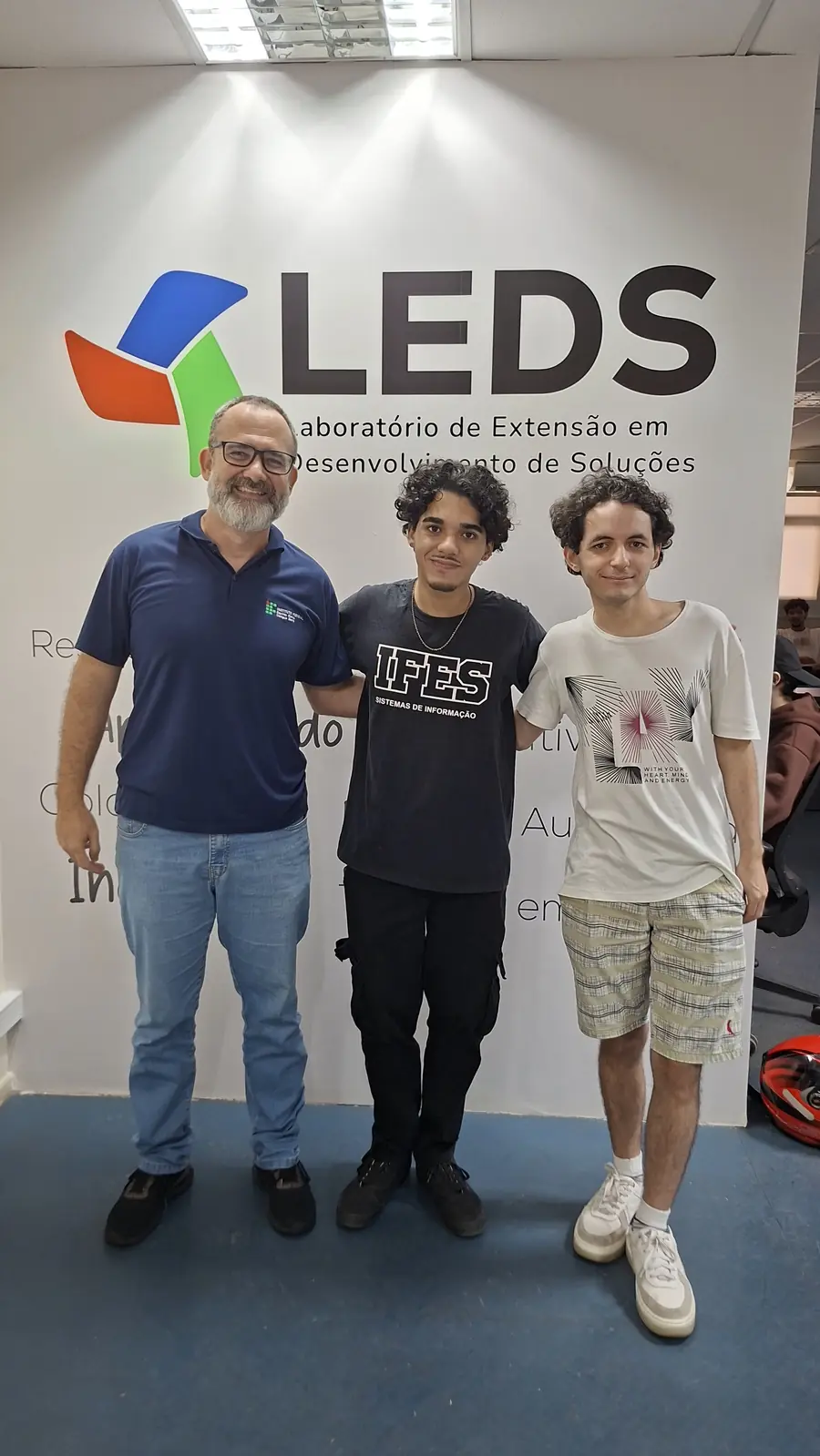 Três integrantes posam diante do painel com a identidade visual do LEDS