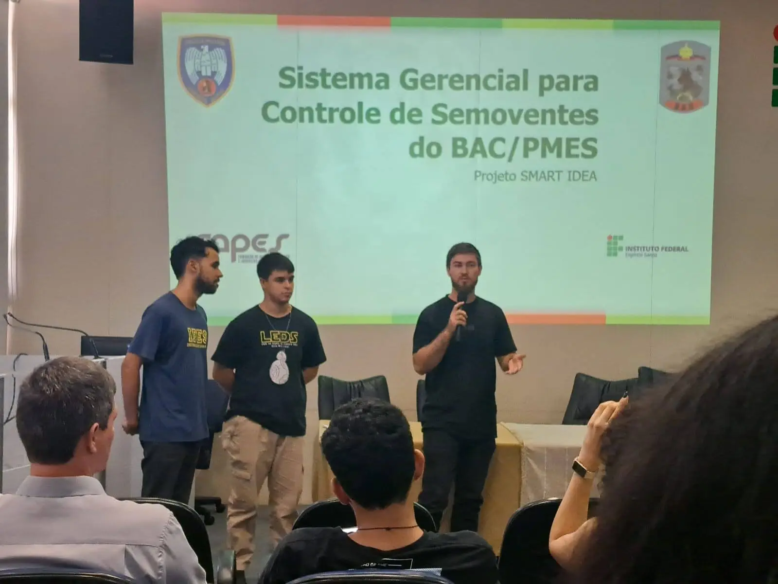 Três integrantes do LEDS apresentam o sistema do BAC/PMES diante do slide com os logos da FAPES e do IFES