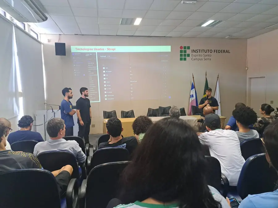 Dois integrantes apresentam no auditório do campus, com as bandeiras ao lado da mesa
