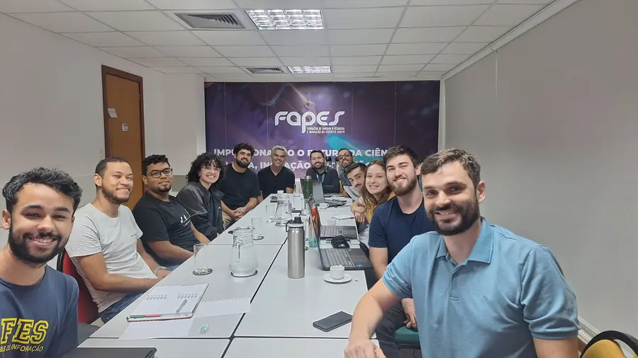 Equipe do LEDS reunida em mesa de reunião, com painel da FAPES ao fundo