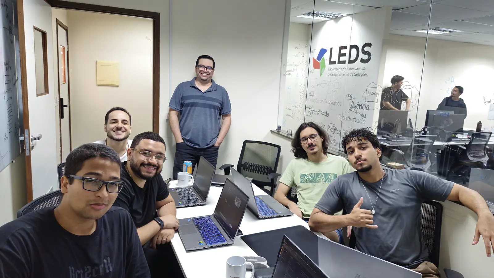 Seis integrantes do LEDS em volta da mesa de trabalho, com a marca do LEDS na divisória de vidro ao fundo