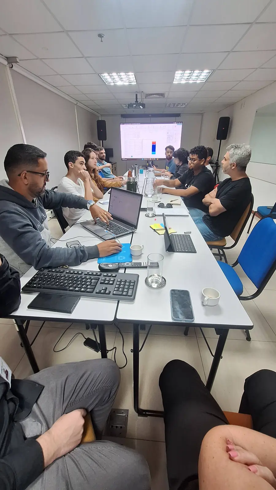 Equipe em volta de mesa comprida com laptops e projetor em sala de reunião