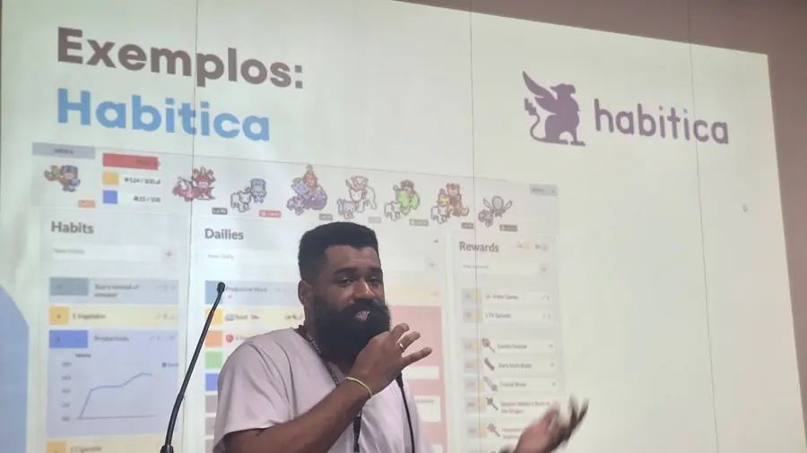 Palestrante fala ao microfone diante do slide 'Exemplos: Habitica'