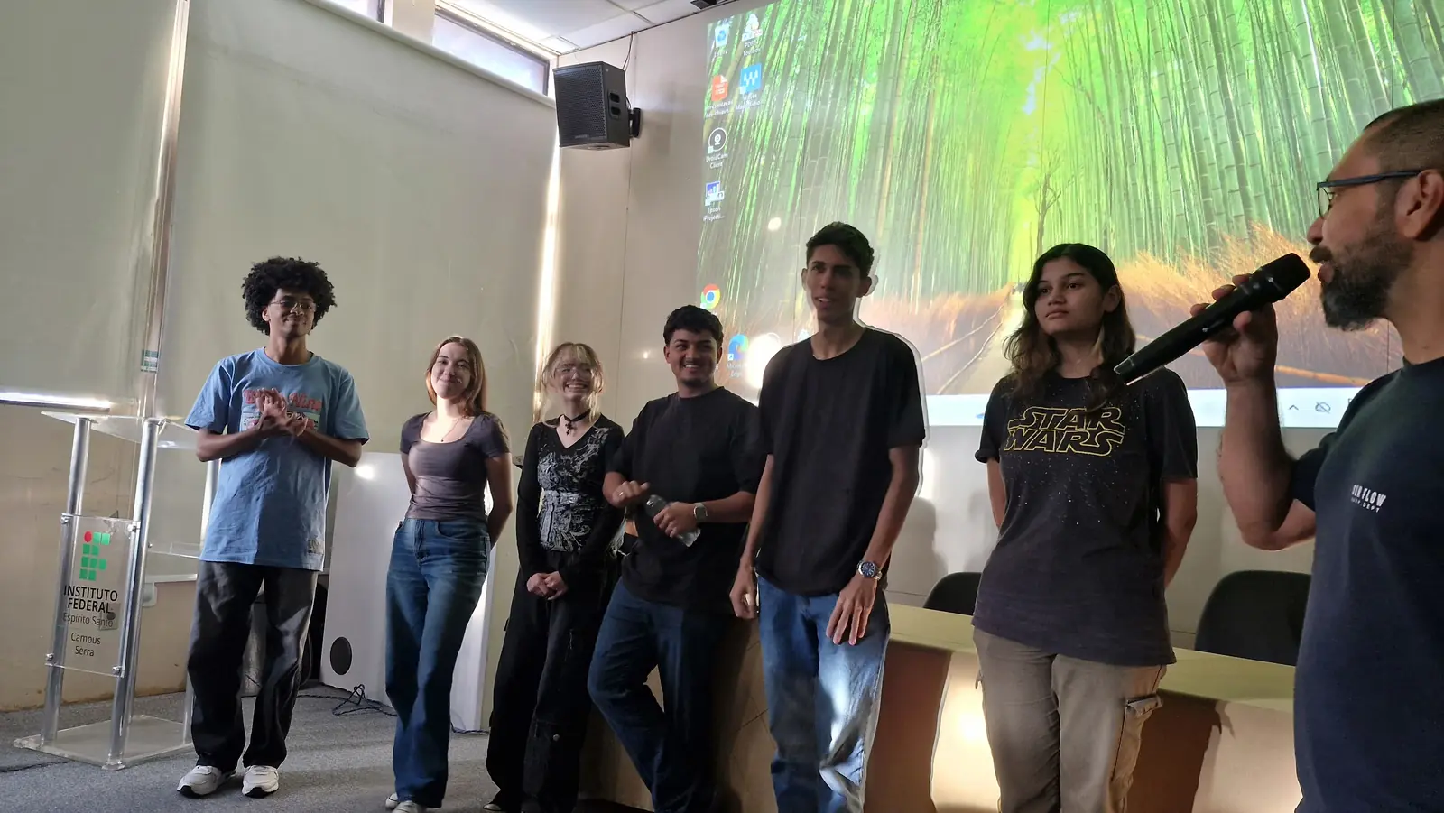 Seis estudantes do Smart Idea em pé no auditório do IFES Campus Serra, ao lado do professor que fala ao microfone